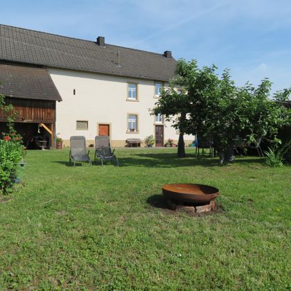 Ein ruhiger Garten mit einer grünen Wiese und einigen Liegestühlen. Im Hintergrund steht ein großes Haus und es gibt eine Feuerstelle.