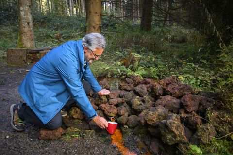 Een man knielt in een bos en giet een vloeistof over een hoop stenen. De omgeving is groen en natuurlijk.