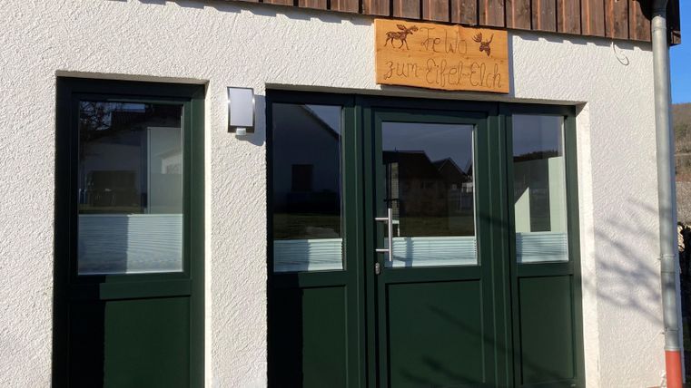 Ein Eingangsbereich mit zwei grünen Türen und einem Holzschild. Die Wände sind hell, und der obere Teil des Gebäudes ist mit Holz verkleidet.