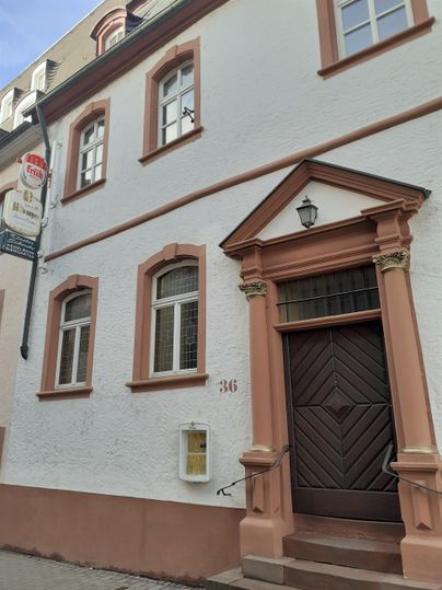 A historic building with a white facade and brown window frames. The entrance door is large and has a striking design.