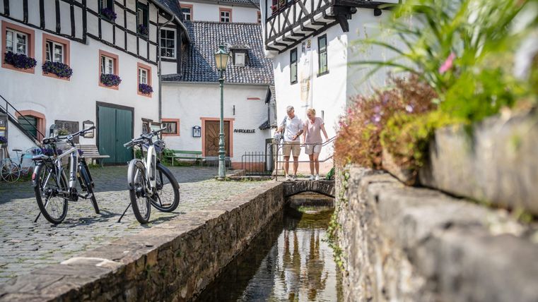 Ein malerischer Platz mit historischen Gebäuden und einem kleinen Bach. Zwei Fahrräder stehen im Vordergrund, während im Hintergrund zwei Personen stehen und die Umgebung genießen.