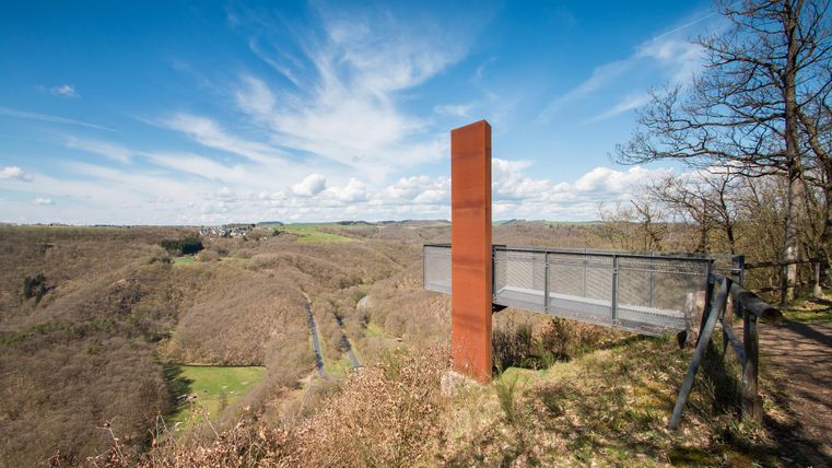 Eine Aussichtsplattform mit einem modernen, roten Kunstwerk blickt über eine hügelige Landschaft. Der Himmel ist klar mit einigen Wolken.