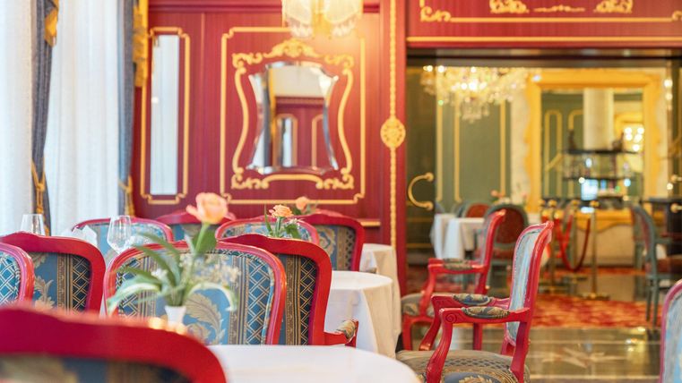 Een elegant restaurant met rode stoelen en decoratieve muren. Grote ramen zorgen voor veel licht en een aangename sfeer.