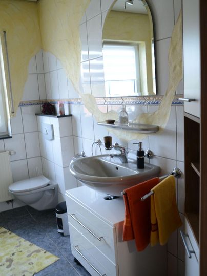 Een moderne badkamer met een wastafel, een grote spiegel en lichte handdoeken. De muren zijn gedeeltelijk geel geschilderd en de tegels zijn grijs.