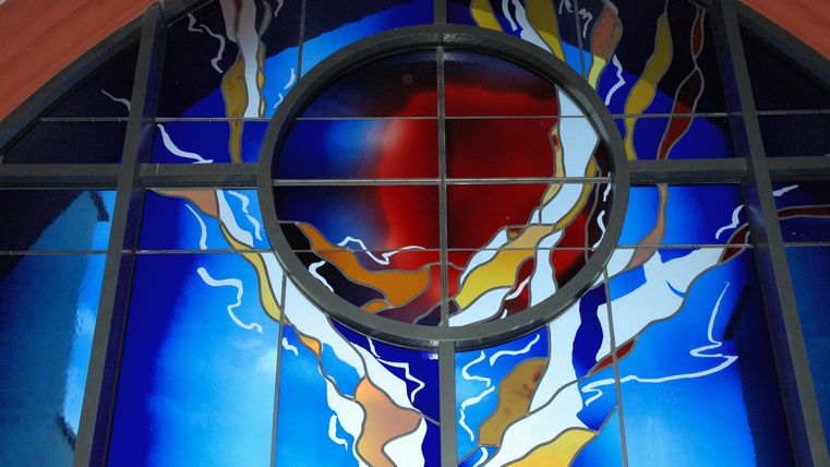 Ein buntes, modernes Glasfenster mit einem großen roten Kreis in der Mitte. Umgeben von verwobenen Linien in Blau, Weiß und Gelb.