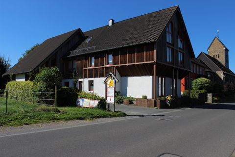 Ein modernes Haus mit Holzverkleidung, umgeben von grünem Rasen und Bäumen. Im Hintergrund ist ein Kirchturm sichtbar und der Himmel ist klar und blau.