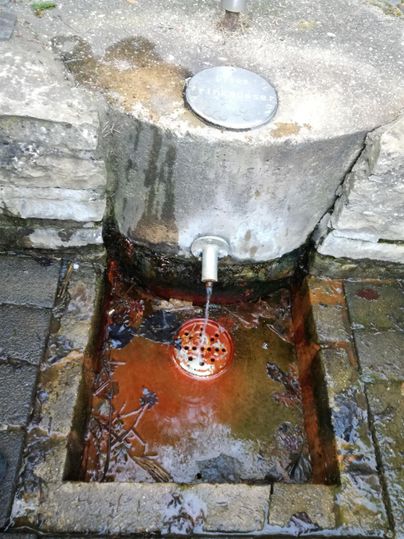 Ein artesischer Brunnen mit rostigem Wasser und einem Gitter. Der Bereich um den Brunnen ist mit Steinen gepflastert.