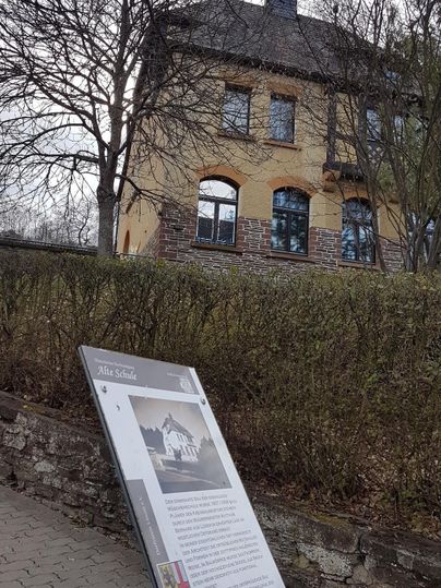 Ein historisches Gebäude steht hinter einer Hecke. Im Vordergrund befindet sich ein Informationstafel über das Gebäude.