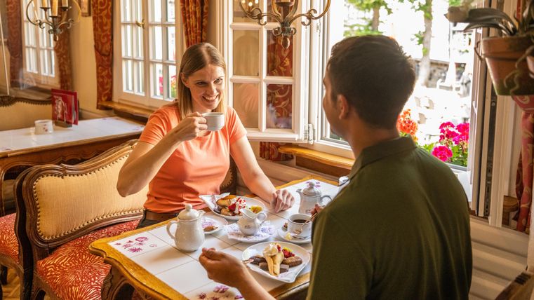 Ein gemütliches Café mit einem Paar, das gemeinsam frühstückt. Sie genießen Kaffee und verschiedene Speisen am Tisch neben einem Fenster mit Blick auf Blumen.