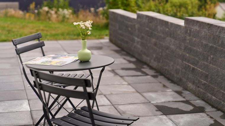 Ein kleiner Tisch mit zwei schwarzen Stühlen steht auf einem gepflasterten Boden. Auf dem Tisch steht eine Vase mit Blumen und eine Zeitschrift.