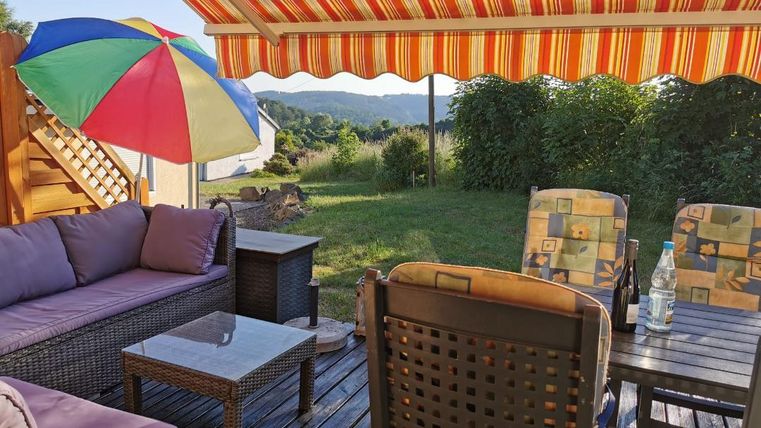 Een gezellige buitenruimte met een bank, een tafel en stoelen. Een kleurrijke parasol en een zonnescherm bieden schaduw, terwijl het groene gras op de achtergrond te zien is.