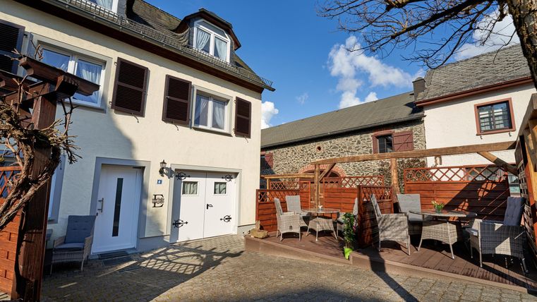 A cozy courtyard with a white house and a terrace. There are garden furniture and a clear blue sky.