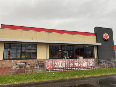 A burger restaurant with a bright facade and a large advertisement banner. The sky is overcast and there are chairs outside.