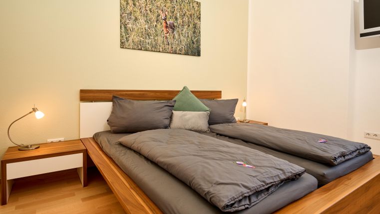 Ein gemütliches Schlafzimmer mit einem Doppelbett und grauer Bettwäsche. An der Wand hängt ein Bild einer Ente, und es gibt einen Nachttisch mit einer Lampe.