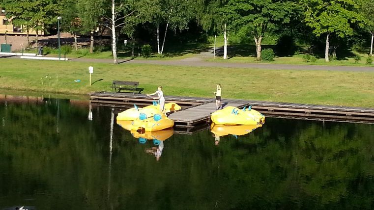 Two yellow pedal boats are moored at a dock on a calm lake. In the background, green trees and a meadow can be seen.
