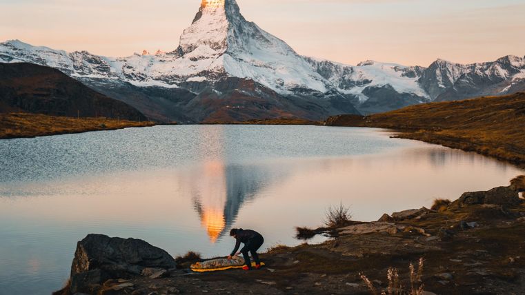 Ein Mann bereitet sich am Ufer eines ruhigen Sees vor, während der majestätische Matterhorn im Hintergrund leuchtet. Die Landschaft ist von Bergen und sanften Hügeln umgeben.