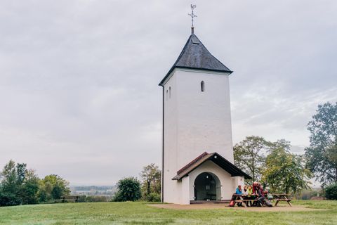 Ein weißer Kirchturm steht in einer grünen Wiese. Im Hintergrund sind Bäume und ein bewölkter Himmel zu sehen.