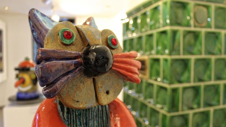 Eine bunte Katzenfigur aus Keramik mit leuchtenden Augen steht vor einer grünen Fliesenwand. Die Statue hat einen einzigartigen, künstlerischen Stil.