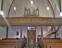 Ein Innenraum einer Kirche mit holzverkleideten Bänken und einer Orgel im Hintergrund. Eine Treppe führt zur Empore.