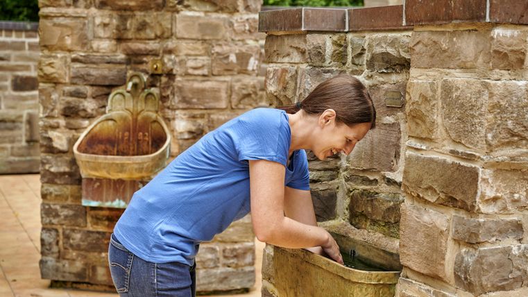 A woman leans over a stone washbasin. In the background, another installation is visible.