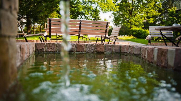 Een rustige tuin met een kleine vijver en stromend water. Op de achtergrond staan houten banken onder grote bomen.