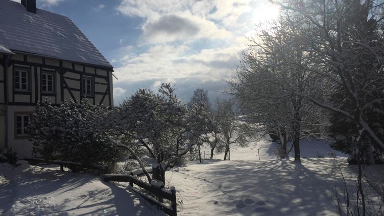 Eine verschneite Landschaft mit einem charmanten Haus und schneebedeckten Bäumen. Der Himmel ist klar mit einigen Wolken und Sonnenstrahlen.