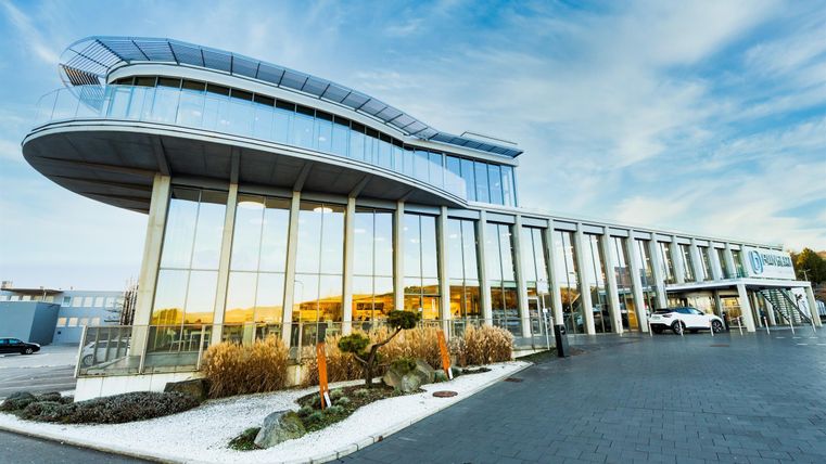 Ein modernes Gebäude mit großen Glasfenstern und einer eleganten Architektur. Die Umgebung ist gepflegt mit Pflanzen und einem klaren Himmel.