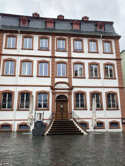 Ein historisches Gebäude mit einer schönen Fassade in Rot und Weiß. Vor dem Eingang stehen Tische mit Sonnenschirmen, und der Platz ist mit Wasser bedeckt.