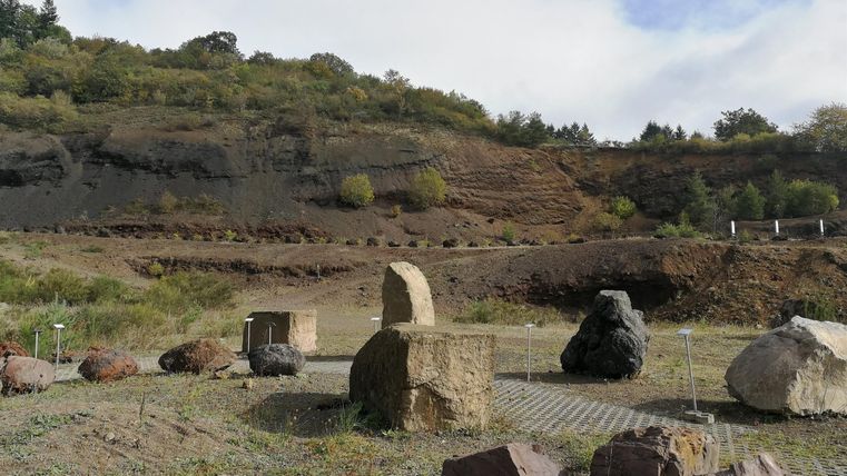 Eine Landschaft mit verschiedenen Steinen und einer bewaldeten Hügelkante im Hintergrund. Der Himmel ist leicht bewölkt.