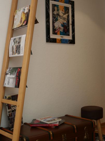 Ein gemütlicher Raum mit einer Holzleiter, auf der Zeitschriften liegen. Neben der Leiter steht ein Koffer und ein Hocker, während ein kunstvolles Bild an der Wand hängt.
