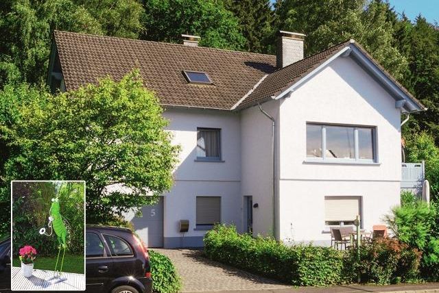 A modern house surrounded by green landscape. In front of the house is a small table with chairs and flowers.