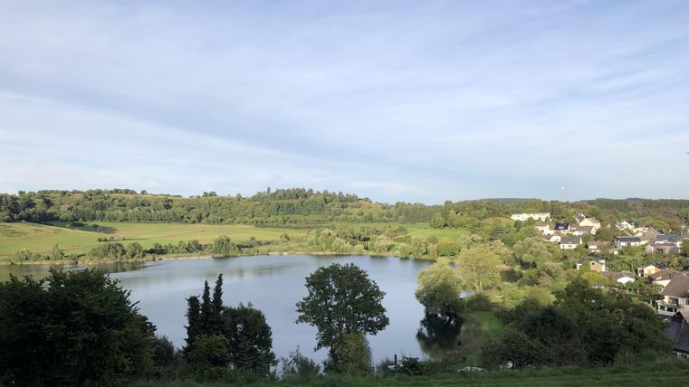 Een rustige meer omgeven door zachte heuvels en bomen. Op de achtergrond zijn enkele huizen en een heldere lucht te zien.