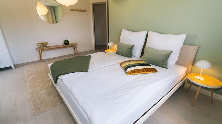 Een moderne slaapkamer met een groot bed en stijlvolle kussens. De muren zijn in zachte groentinten en er is een gezellige sfeer met minimalistische inrichting.