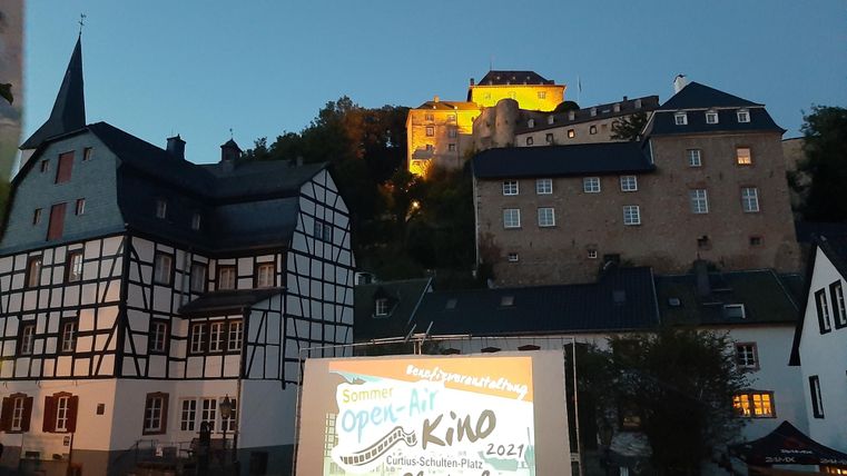 Een schilderachtige plek met historische gebouwen en een groot scherm voor een openluchtbioscoop. Op de achtergrond straalt een kasteel in de schemering.