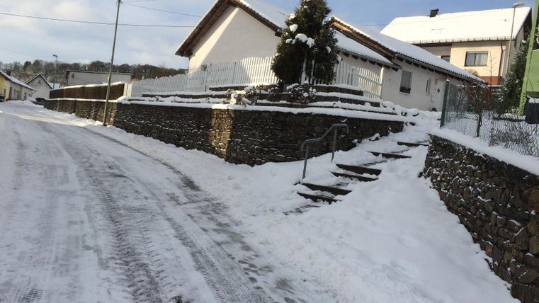 Eine verschneite Straße führt zu einem Haus. Der Weg ist von Schnee bedeckt und die Umgebung wirkt ruhig und winterlich.