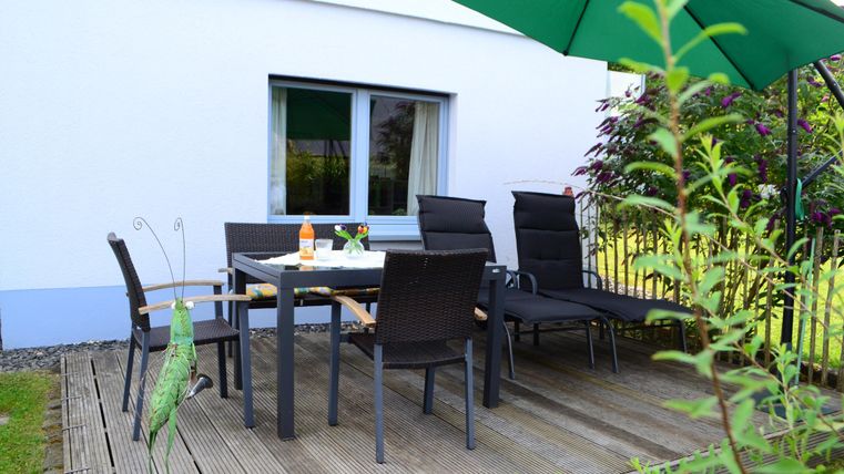 A cozy outdoor area with a table and chairs on a wooden terrace. Above the table is a green sunshade, and nearby plants are growing.