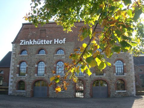 A historic building named Zinkhütter Hof with large windows and a stone facade. In the foreground, green leaves can be seen.