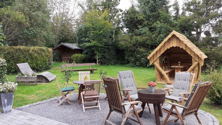 Ein gemütlicher Garten mit Sitzgelegenheiten und einer kleinen Holzlaube. Im Hintergrund sind Bäume und ein weiterer Holzschuppen zu sehen.