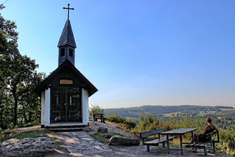 Eine kleine Kapelle mit einem Kreuz steht auf einem Hügel. Im Hintergrund erstreckt sich eine malerische Landschaft unter einem klaren blauen Himmel.