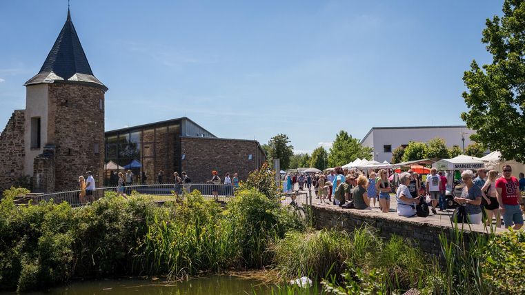 A gathering of people stands by a waterway in front of a historic building with a tower-like roof. The surroundings are green and sunny, perfect for outdoor activities.