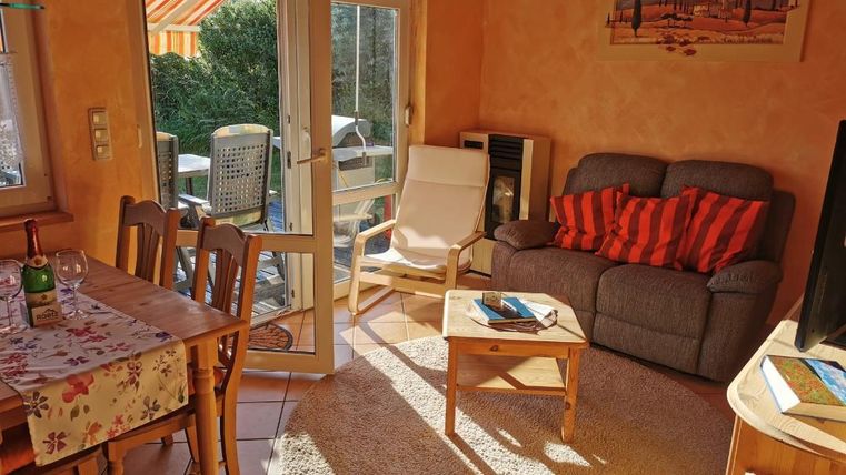 Een gezellige woonkamer met een grijze bank en een kleine tafel. Er is een houten eettafel en veel daglicht dat door de ramen stroomt.