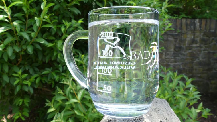 Ein klarer Glasbecher steht auf einem Stein, umgeben von grünen Pflanzen. Der Becher hat eine Markierung und eine Gravur mit der Aufschrift "BAD".