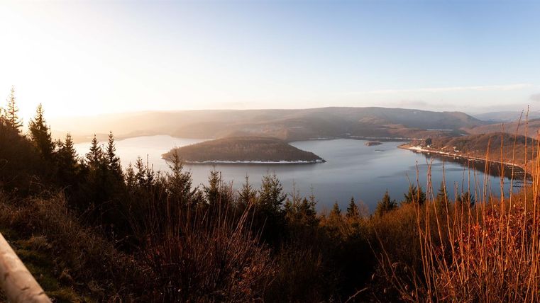Eine malerische Aussicht auf einen See, umgeben von sanften Hügeln und Wäldern. Der Himmel leuchtet in warmen Farben der Abenddämmerung.