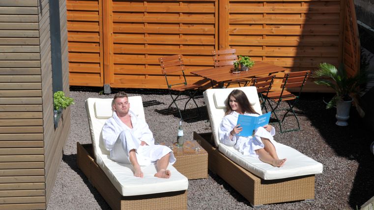 Ein entspannender Außenbereich mit zwei Liegen. Ein Mann und eine Frau in Bademänteln genießen die Sonne und lesen.