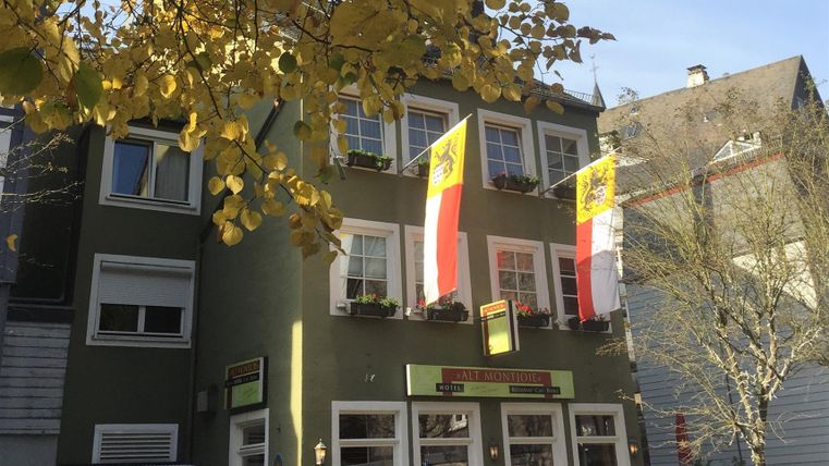 Ein grünes Gebäude mit großen Fenstern und zwei Fahnen. Im Vordergrund sind Passanten und herbstliche Blätter zu sehen.