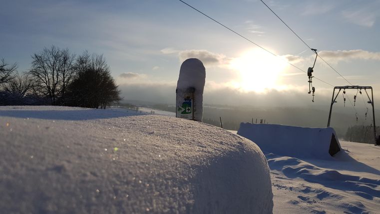 Eine verschneite Landschaft mit einem Sonnenaufgang im Hintergrund. Im Vordergrund liegt eine dicke Schicht Schnee und spielende Schaukel.