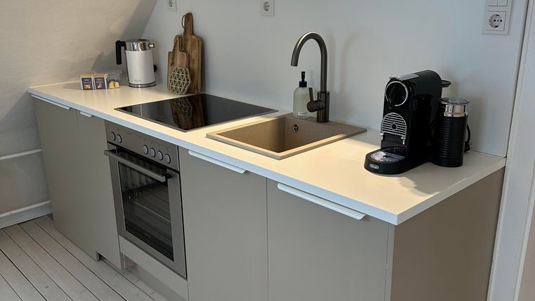 Een moderne keuken met lichte kasten, een roestvrijstalen fornuis en een gootsteen. Op het werkblad staan een koffiezetapparaat en decoratieve elementen.