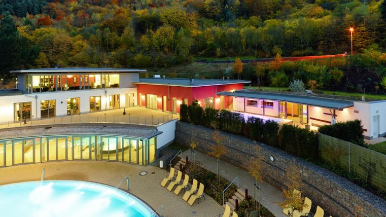 Een modern wellnessresort met een zwembad en ligstoelen op de voorgrond. Op de achtergrond zijn de kleurrijke bomen van het landschap te zien.
