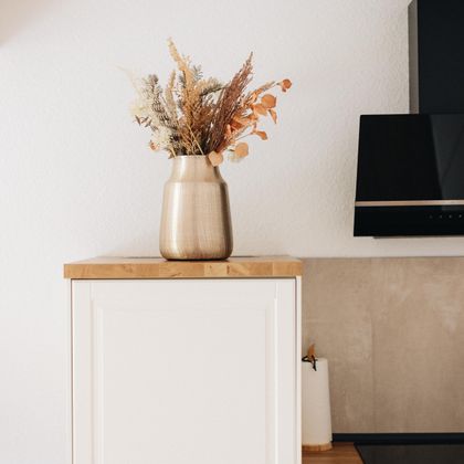 Eine moderne Küche mit einem weißen Kühlschrank und einer Holzarbeitsplatte. Auf dem Kühlschrank steht eine Vase mit Trockenblumen.