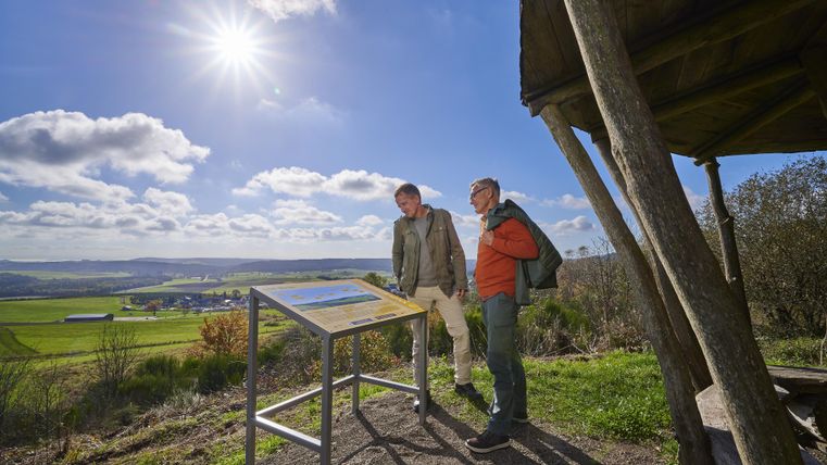 Twee personen staan bij een informatieschild in de natuur. Op de achtergrond ziet men een groene landschap onder een heldere lucht.